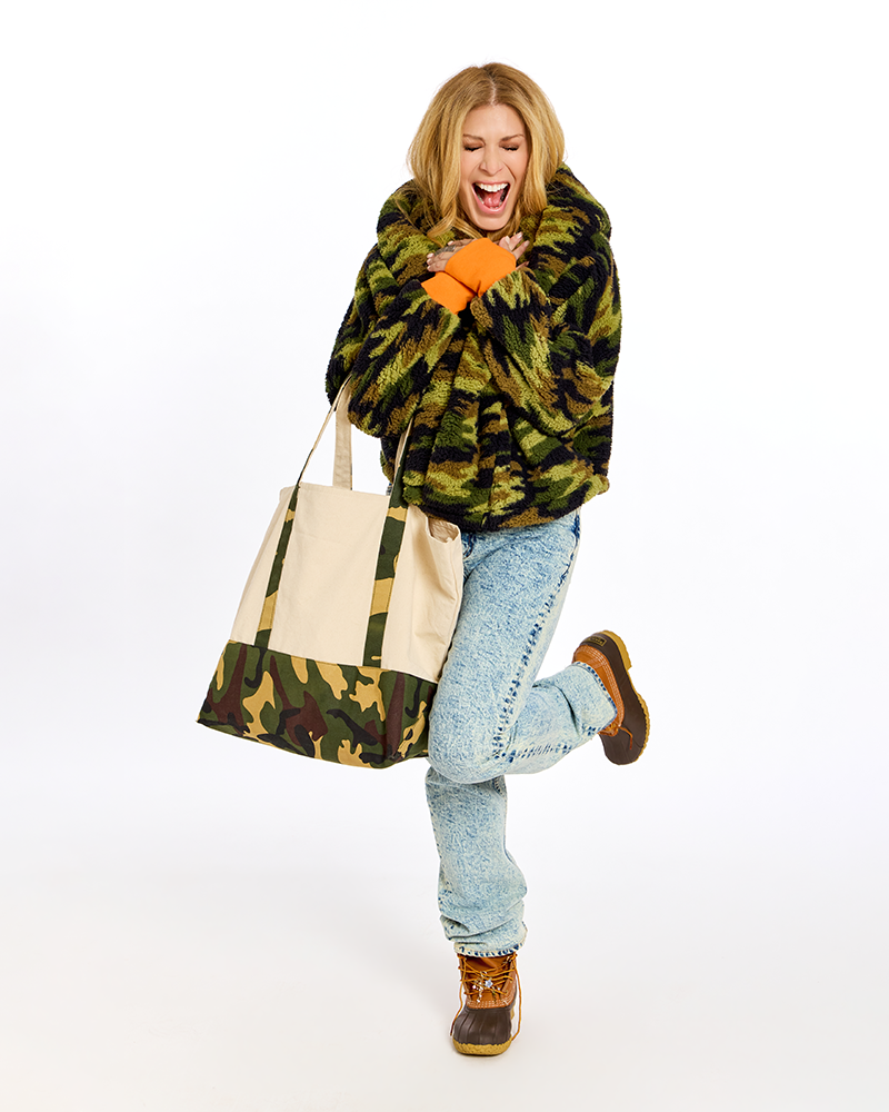 A woman in a green and black camouflage faux fur coat, light blue jeans, and brown boots is holding a large canvas tote bag with a matching camouflage bottom panel, set against a plain white background.|Jill is 5'6" wearing XS/S