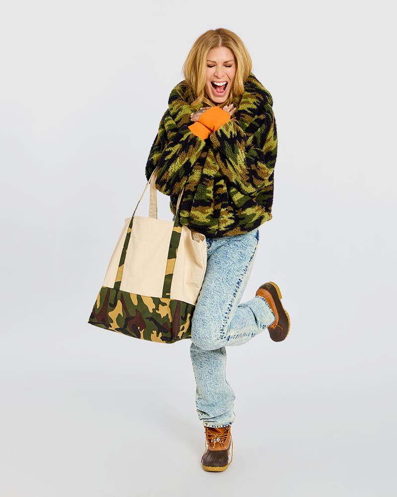 A woman in a green and black camouflage faux fur coat, light blue jeans, and brown boots is holding a large canvas tote bag with a matching camouflage bottom panel, set against a plain white background.|Jill is 5'6" wearing size XS/S