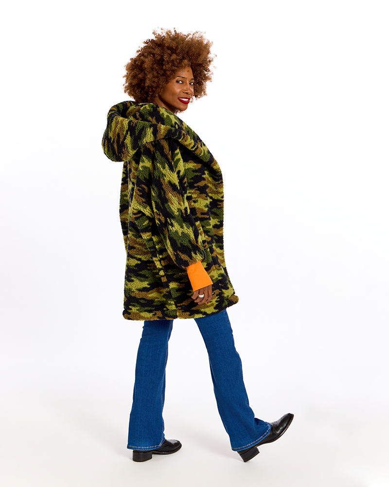 A woman with an afro hairstyle models a green and black camouflage hooded jacket, paired with blue jeans and black shoes, against a plain white background.