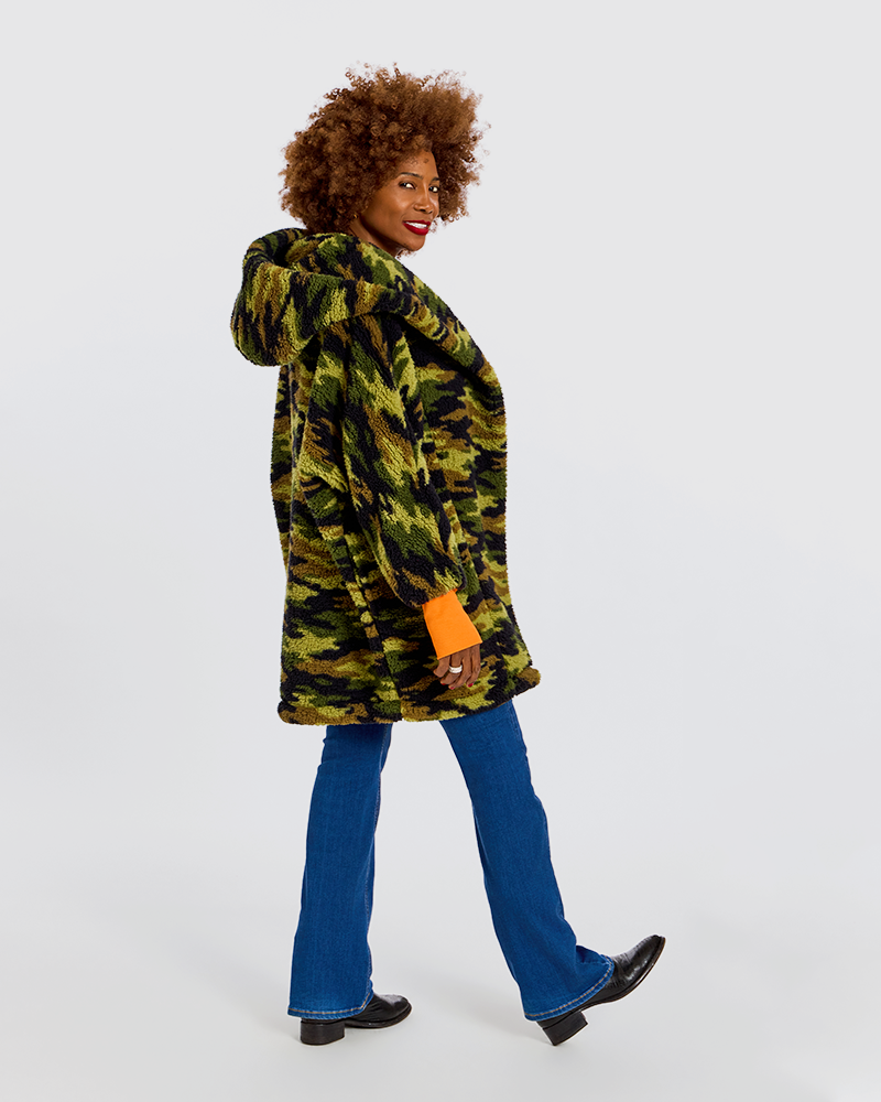 A woman with an afro hairstyle models a green and black camouflage hooded jacket, paired with blue jeans and black shoes, against a plain white background.|Model is 5'7" wearing size XS/S
