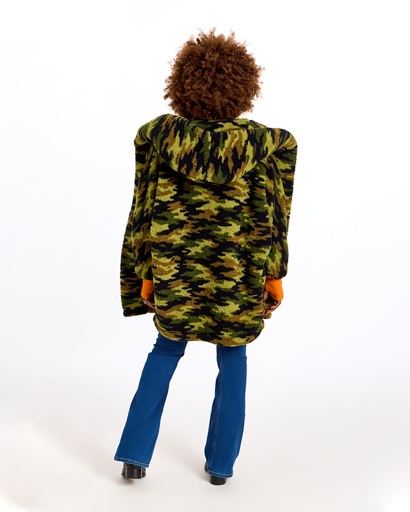 A person shown from the back wearing a green and black camouflage hooded jacket, blue flared jeans, and black shoes, photographed against a plain white studio background.