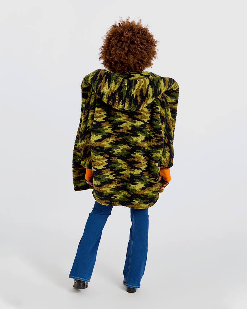 A person shown from the back wearing a green and black camouflage hooded jacket, blue jeans, and orange gloves, standing against a plain light gray studio background.|Model is 5'7" wearing size XS/S