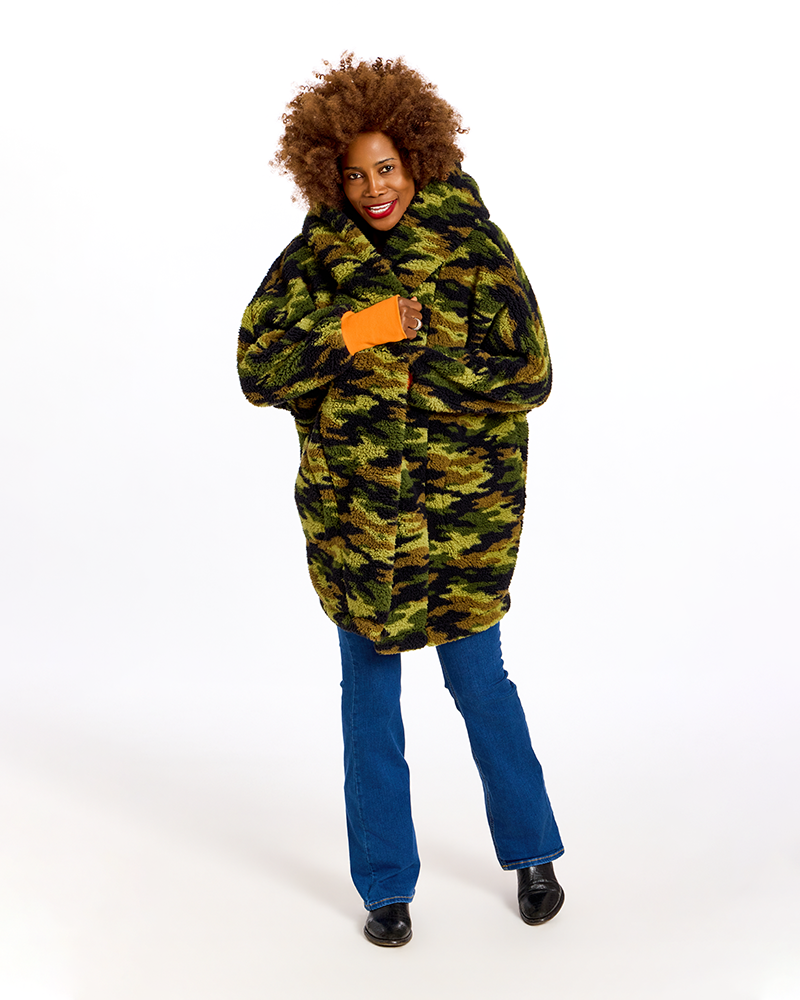 A woman with an afro hairstyle models a long camouflage-patterned faux fur coat with a hood, paired with blue jeans and black shoes, against a solid white studio background.