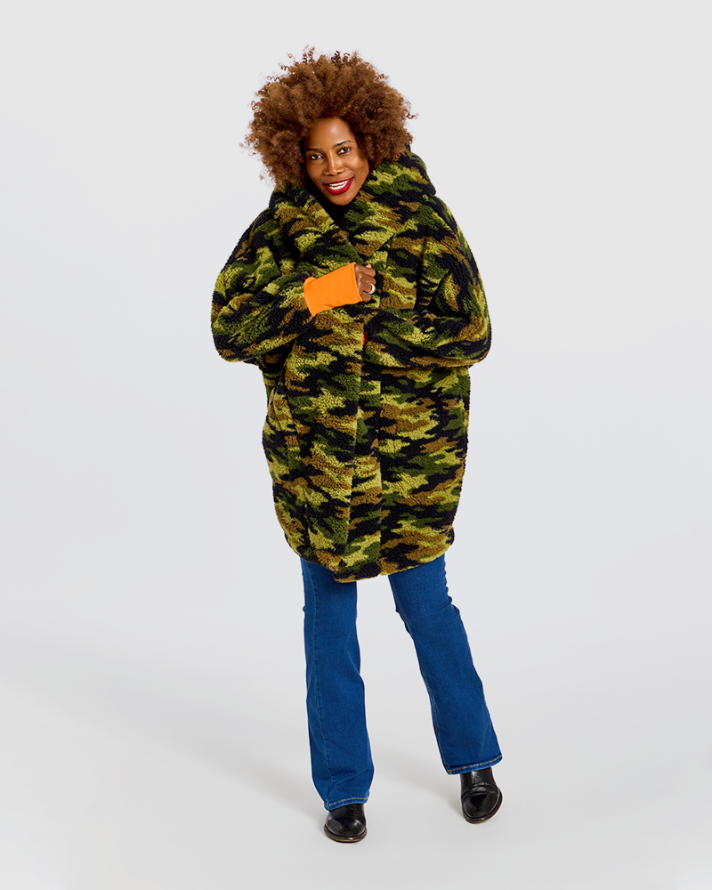 A woman modeling a hooded camouflage puffer jacket with a bright orange inner lining, paired with blue jeans and black shoes, against a plain light gray studio background.|Model is 5'7" wearing size XS/S