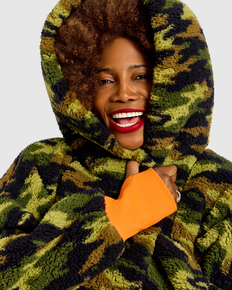 A smiling woman wearing a plush hooded jacket in a green, olive, and black camouflage pattern, with a bright orange cuff detail on the sleeve, against a light gray background.