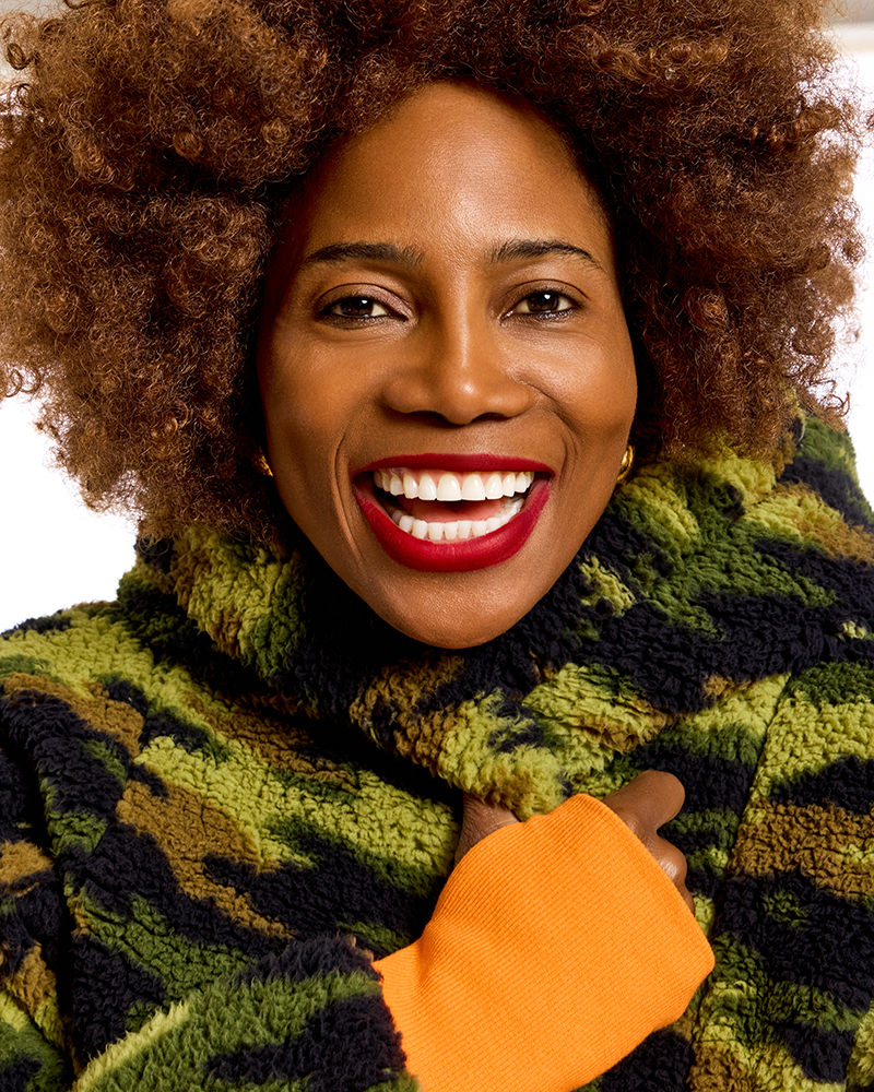 A close-up portrait of a smiling woman with voluminous curly brown hair, wearing a green and black textured fleece jacket with orange cuffs, set against a plain white background.