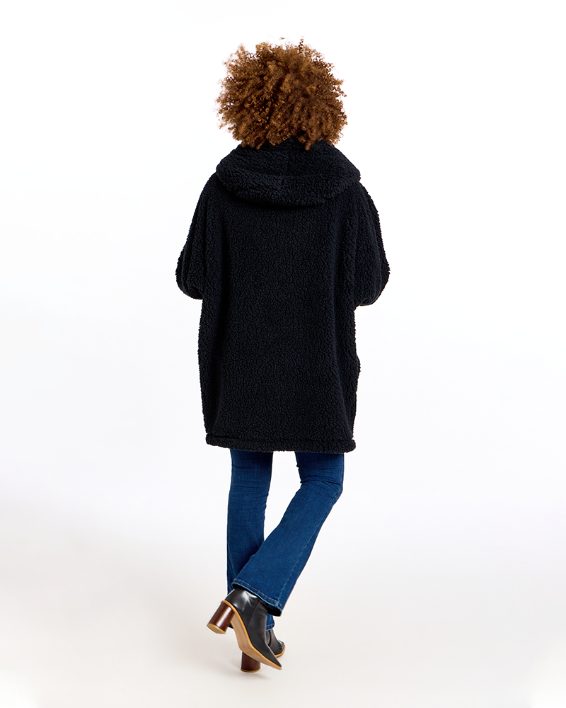 A person shown from the back wearing a black teddy fleece hooded cardigan, blue jeans, and brown and black heeled boots against a plain white studio background.