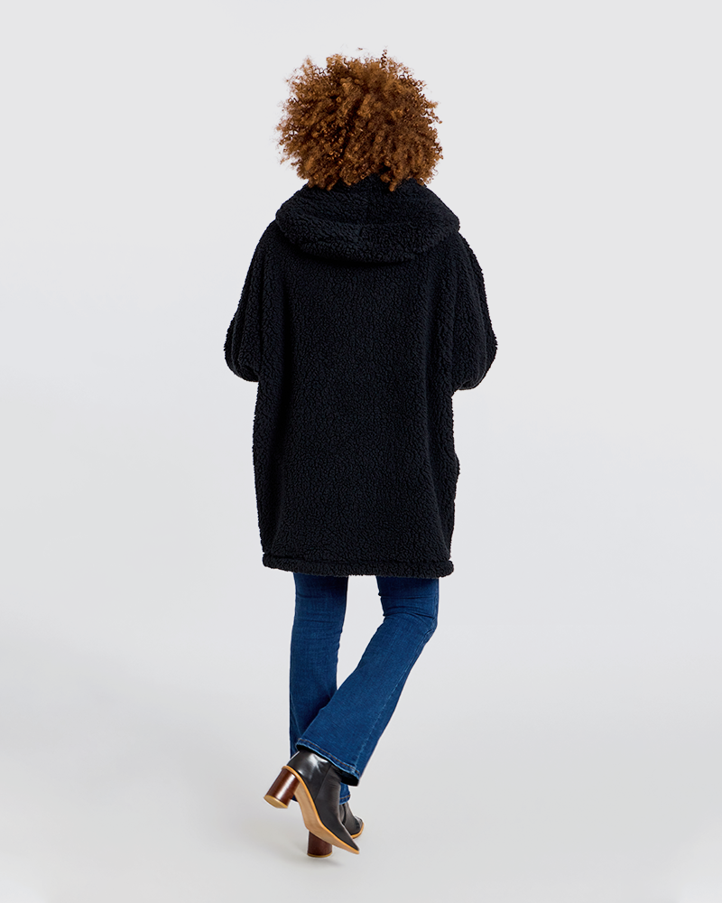 A person shown from the back wearing a black teddy fleece hooded jacket, blue jeans, and brown-and-black heeled boots against a plain light gray studio background.|Model is 5'7" wearing size XS/S