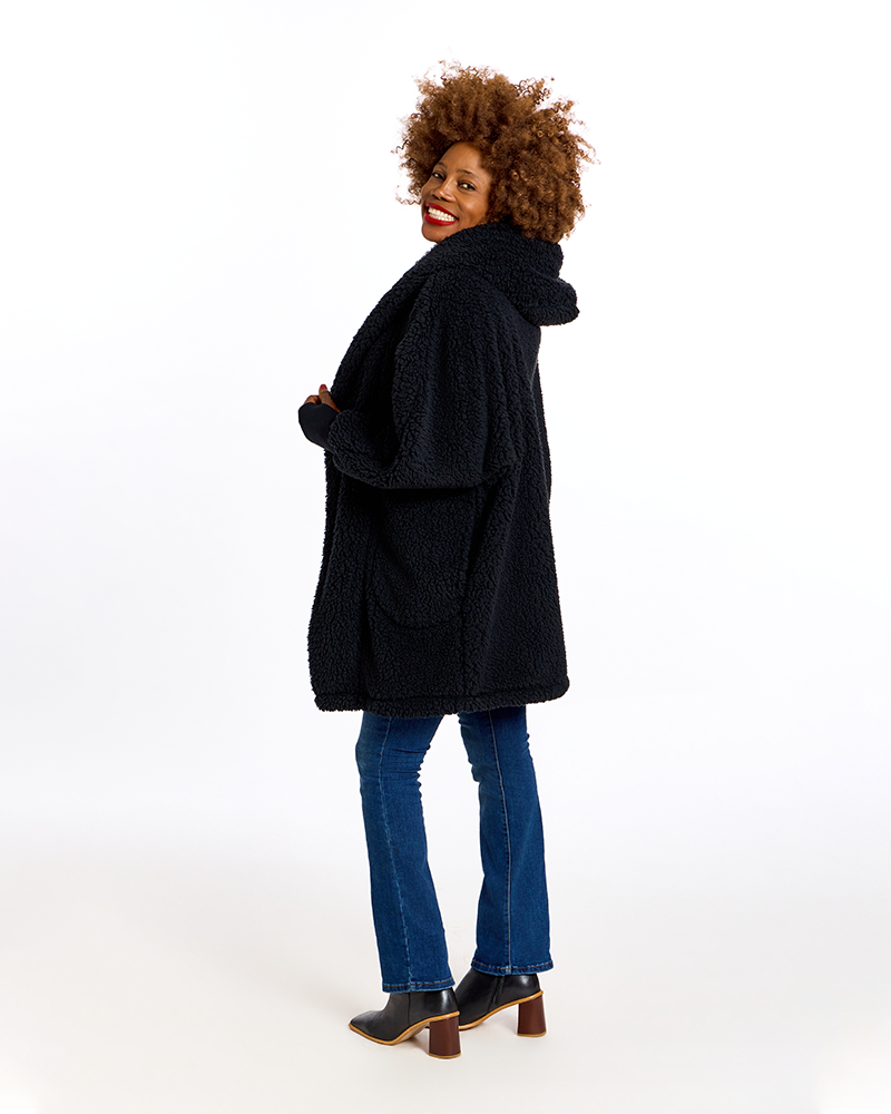 A woman with curly brown hair models a black sherpa fleece hooded coat, paired with blue jeans and black boots, against a solid white studio background.