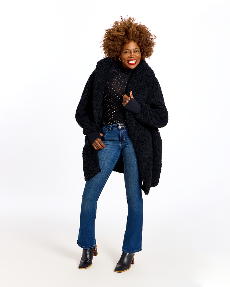 A woman modeling a black teddy coat over a black polka-dot top, blue jeans, and black ankle boots against a plain white studio background.