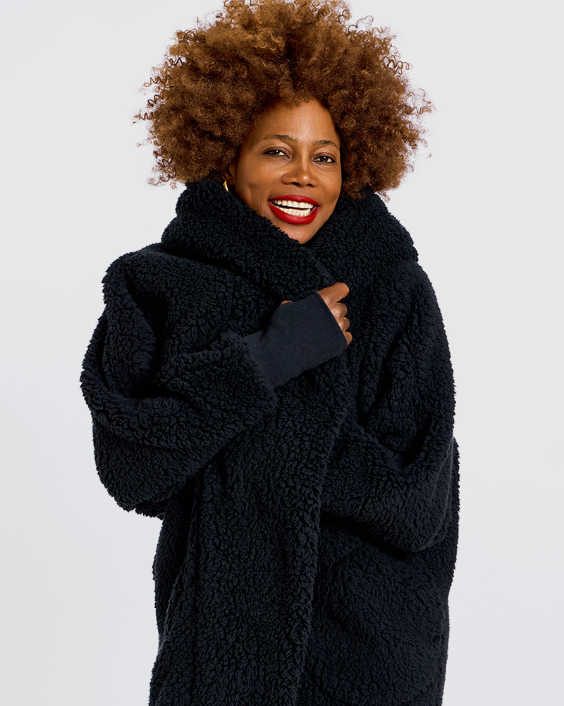 A smiling woman with an afro hairstyle wearing a thick, dark navy blue sherpa fleece coat and matching scarf, photographed against a solid light gray background.|Model is 5'7" wearing size XS/S