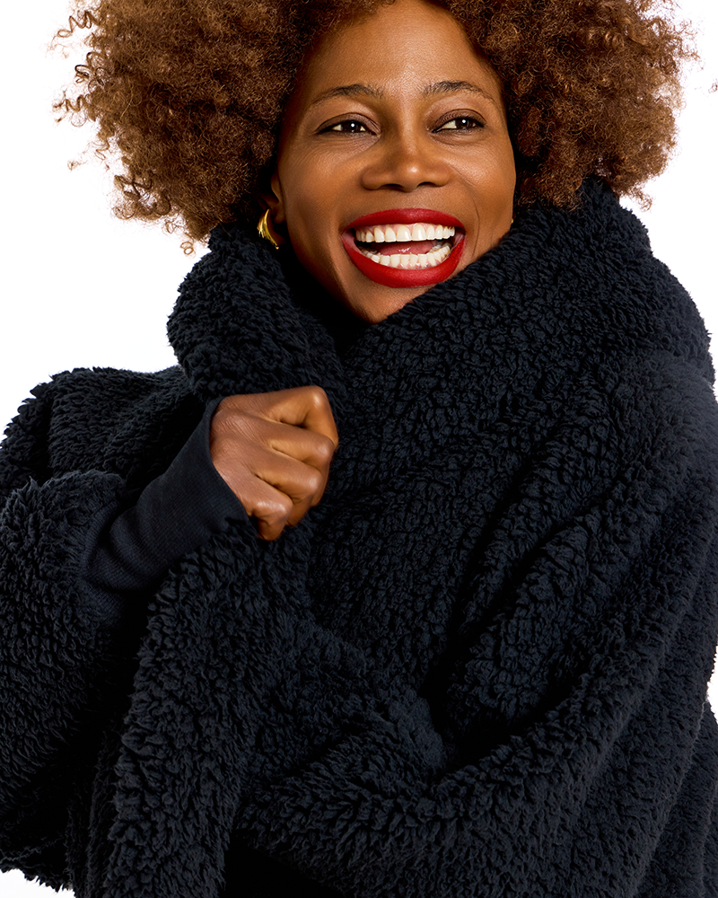 A smiling woman with curly hair wearing a thick, plush black sherpa fleece wrap or coat against a plain white background.