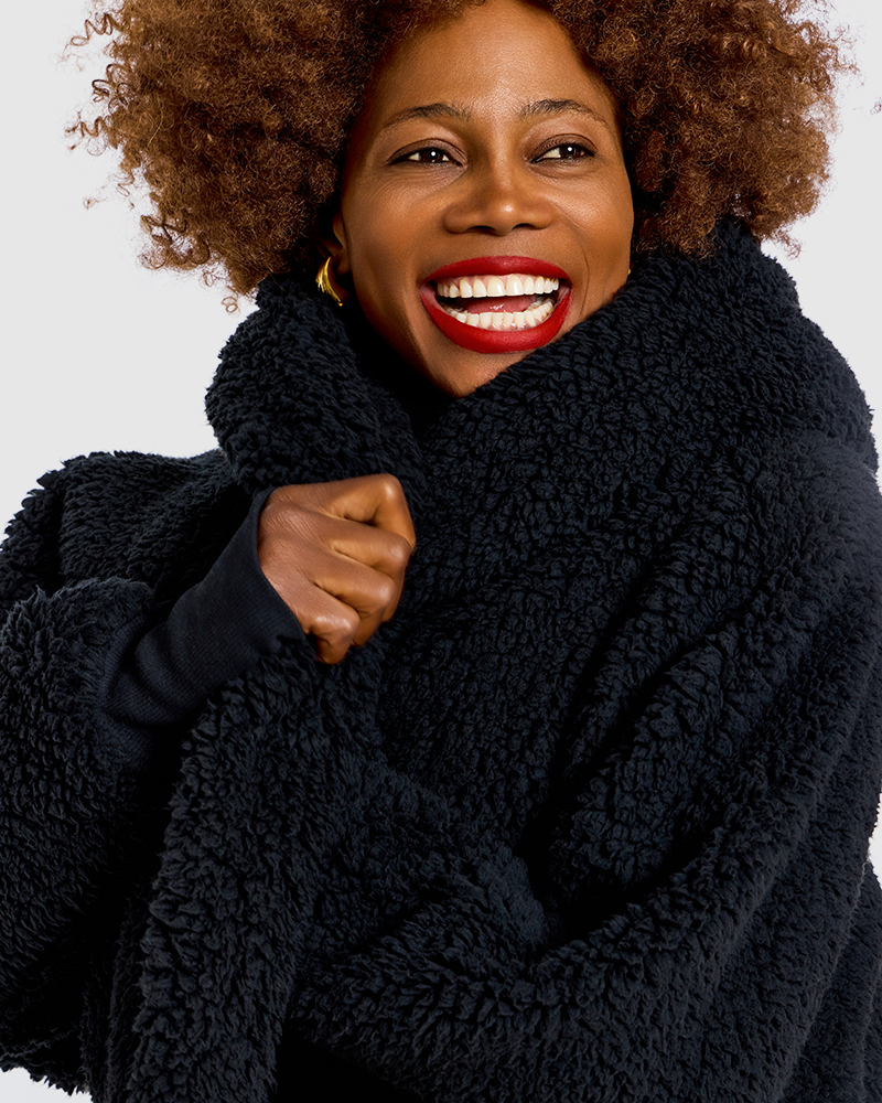 A smiling woman with curly hair wearing a thick, plush black sherpa fleece wrap or coat against a plain white background.|Model is 5'7" wearing size XS/S