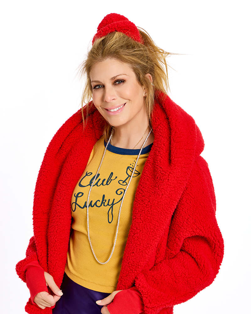 A woman modeling a bright red sherpa fleece hooded jacket and matching red knit beanie against a plain white studio background. She is also wearing a mustard yellow graphic T-shirt and dark purple pants.|Jill: 5'6" in size XS/S