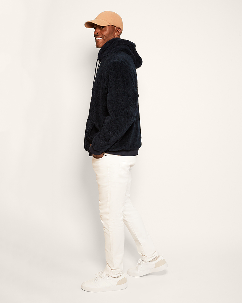 Man wearing a navy hoodie, beige cap, and white pants on a light gray background