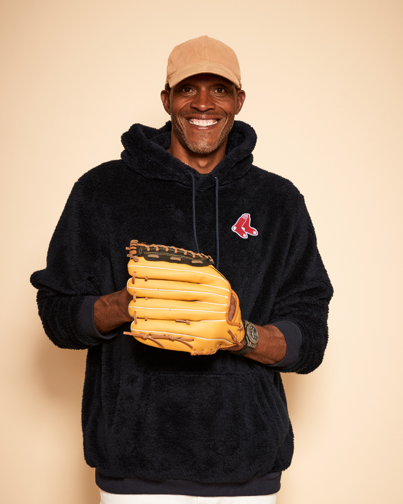 Man wearing a navy hoodie with red sox logo, holding a yellow baseball glove against a beige background