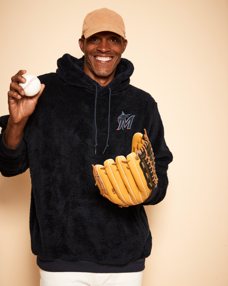 Person wearing a black hoodie with a logo, holding a baseball and a yellow glove against a beige background