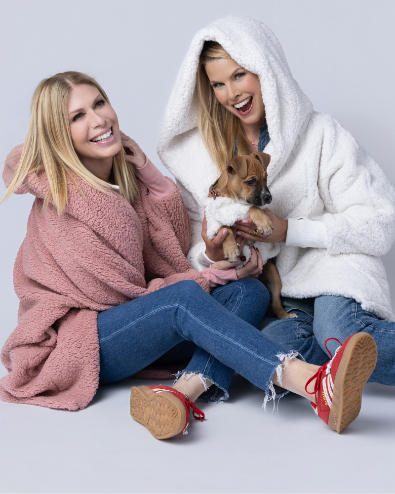 Two women wrapped in cozy blankets with a dog wearing sherpa pet jacket on a grey background