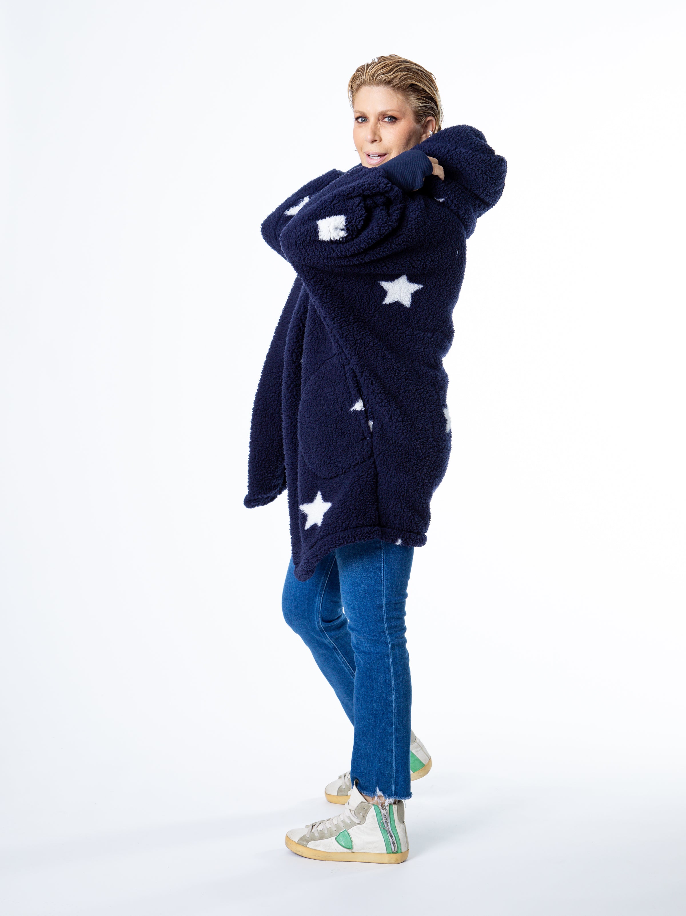 Wearing the Classic Lounger by Jill Martin, a person poses playfully in a dark blue fuzzy coat with white star patterns. They stand sideways, smiling with one hand inside the collar, paired with jeans and white sneakers featuring green accents against a plain white backdrop.