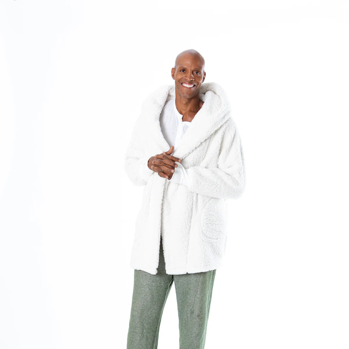 A happy individual models the Limited Edition Penn State White Out Lounger by Jill Martin, an oversized cozy white fluffy jacket with the hood up, paired with green pants against a plain white backdrop.