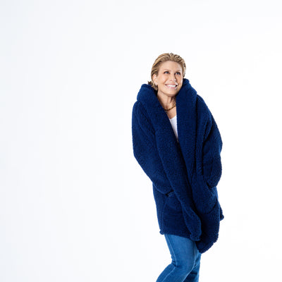 Sporting a Jill Martin Classic Lounger in navy, with a white top and blue jeans, the person stands cross-legged with hands in pockets. They smile subtly while gazing aside, set against a plain white studio backdrop.