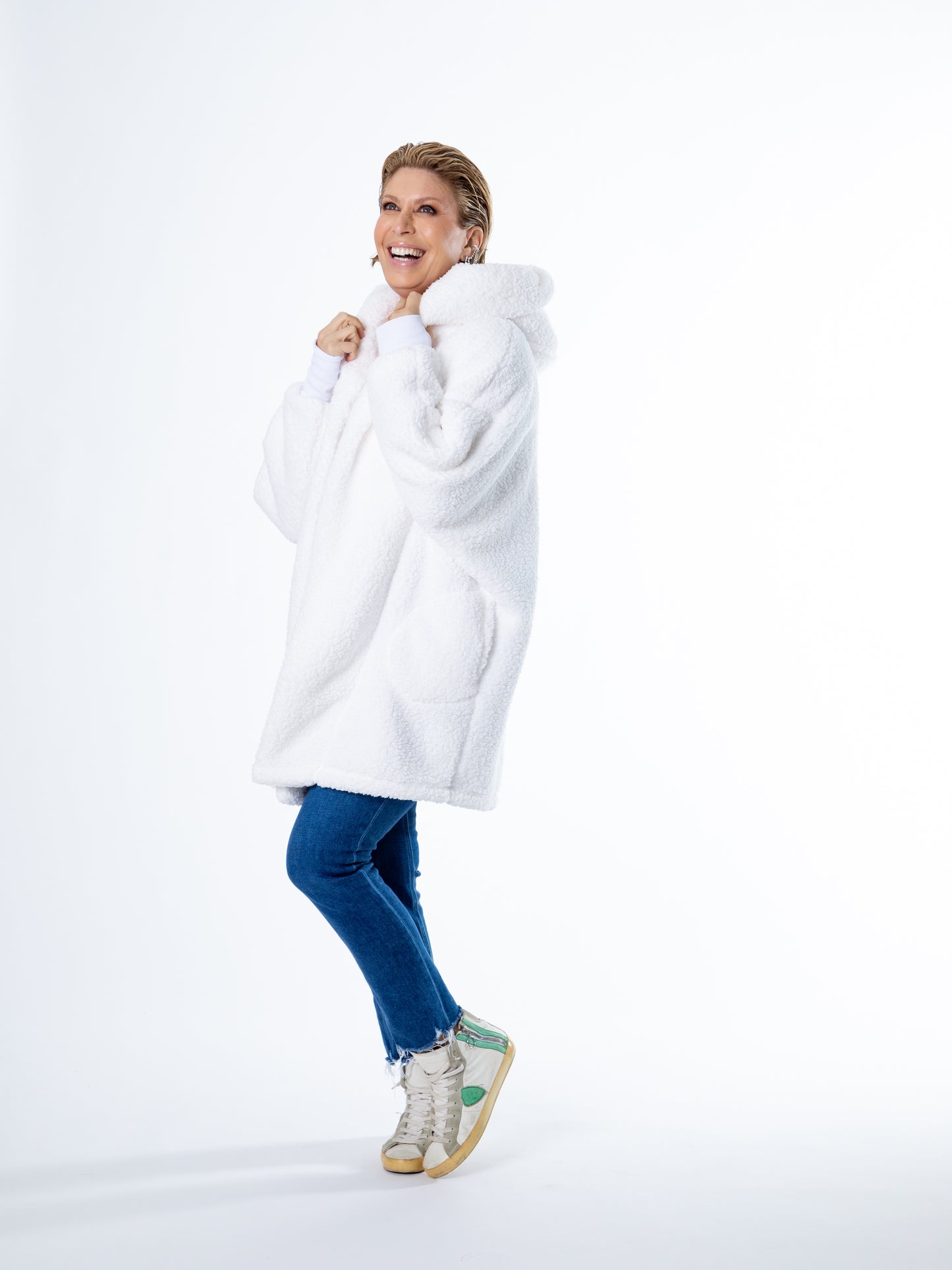 A person grins while wearing the Jill Martin Limited Edition Penn State White Out Lounger, a large cozy fleece hoodie with a hood. They pair it with blue jeans and white sneakers featuring green accents, standing slightly bent at one knee against a plain white backdrop.