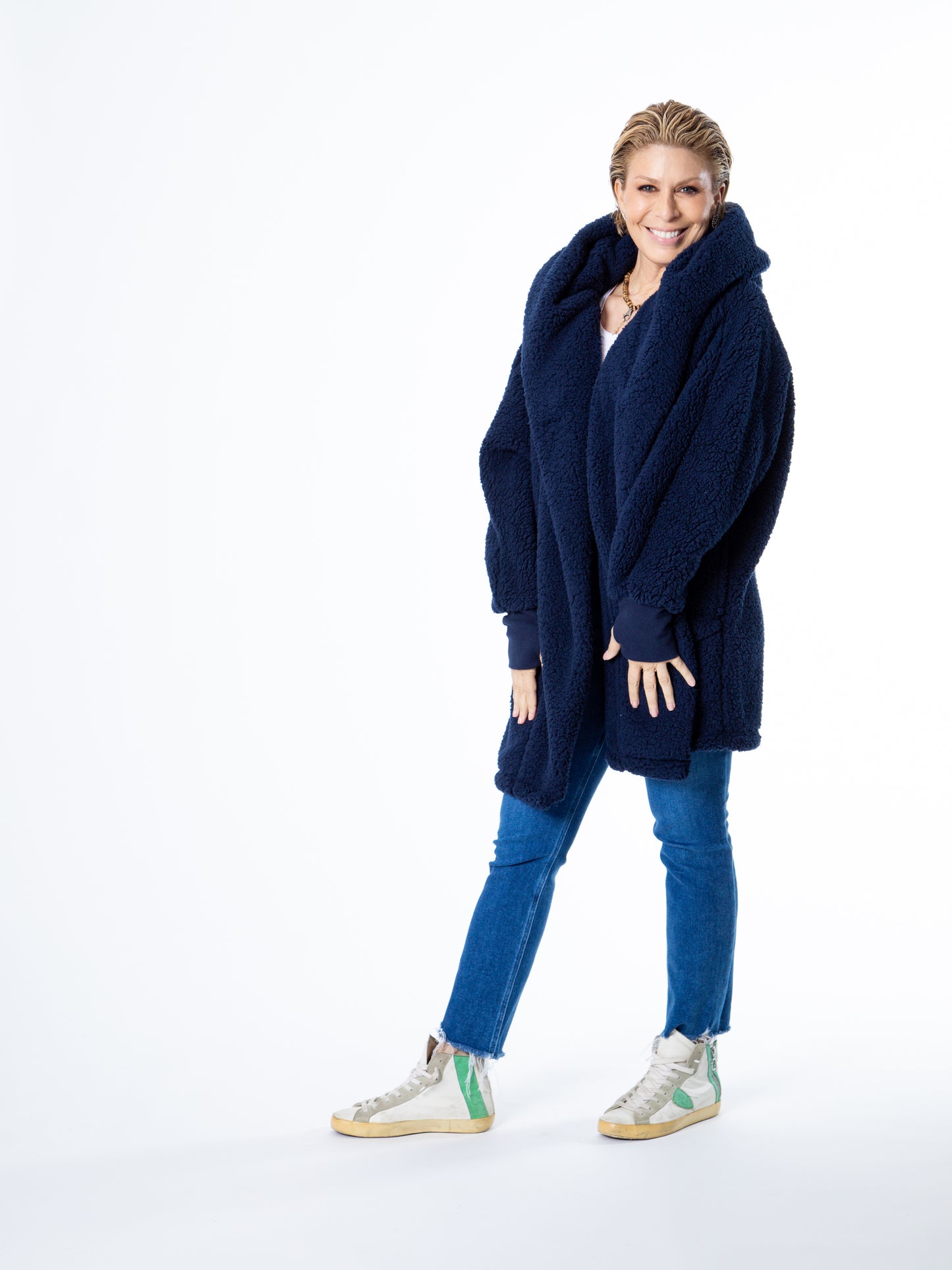 Smiling person with short blonde hair wears the Jill Martin University of Michigan Lounger, blue jeans, and white high-top sneakers with green detailing. They stand against a plain white background, holding the edges of their coat.