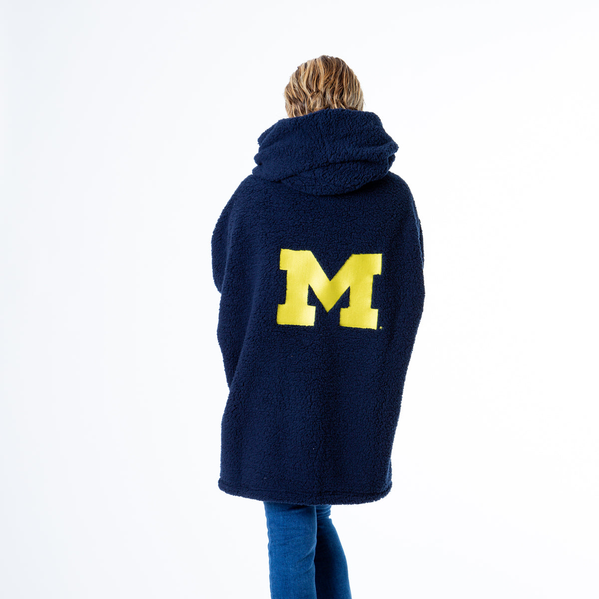 A person stands with their back to the camera in a University of Michigan Lounger by Jill Martin, featuring a large navy M logo. Theyre also wearing blue jeans and light-colored shoes.