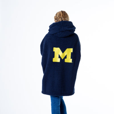 A person stands with their back to the camera in a University of Michigan Lounger by Jill Martin, featuring a large navy M logo. Theyre also wearing blue jeans and light-colored shoes.