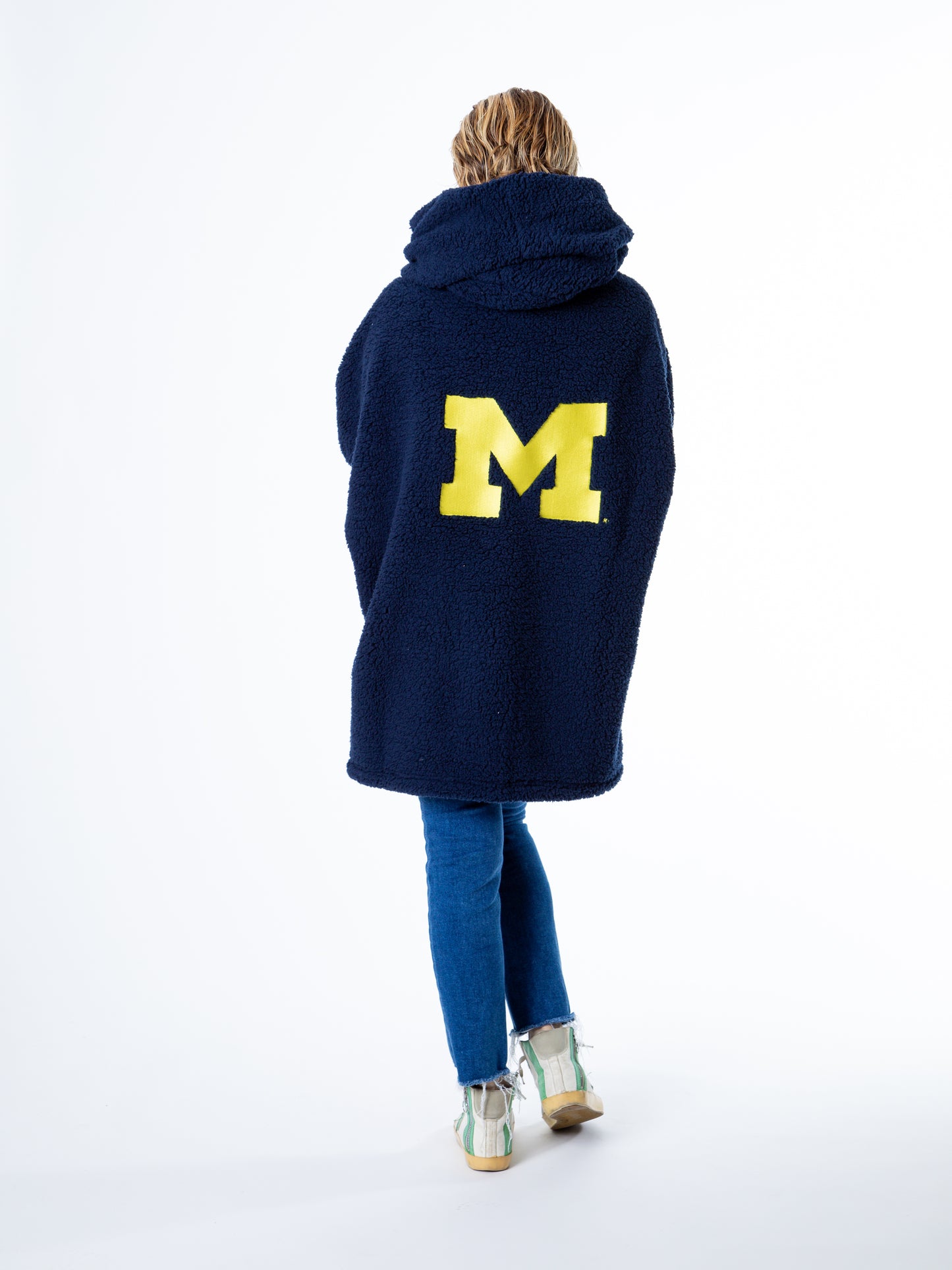 A person stands with their back to the camera in a University of Michigan Lounger by Jill Martin, featuring a large navy M logo. Theyre also wearing blue jeans and light-colored shoes.